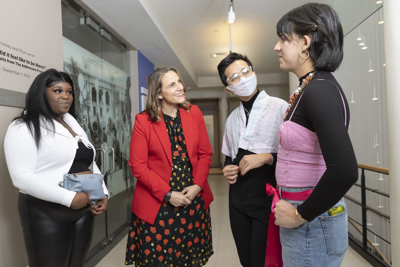 Three people standing and listening to a fourth person on the right speak in the hallway at The Center with archival photography of a protest hanging behind them.