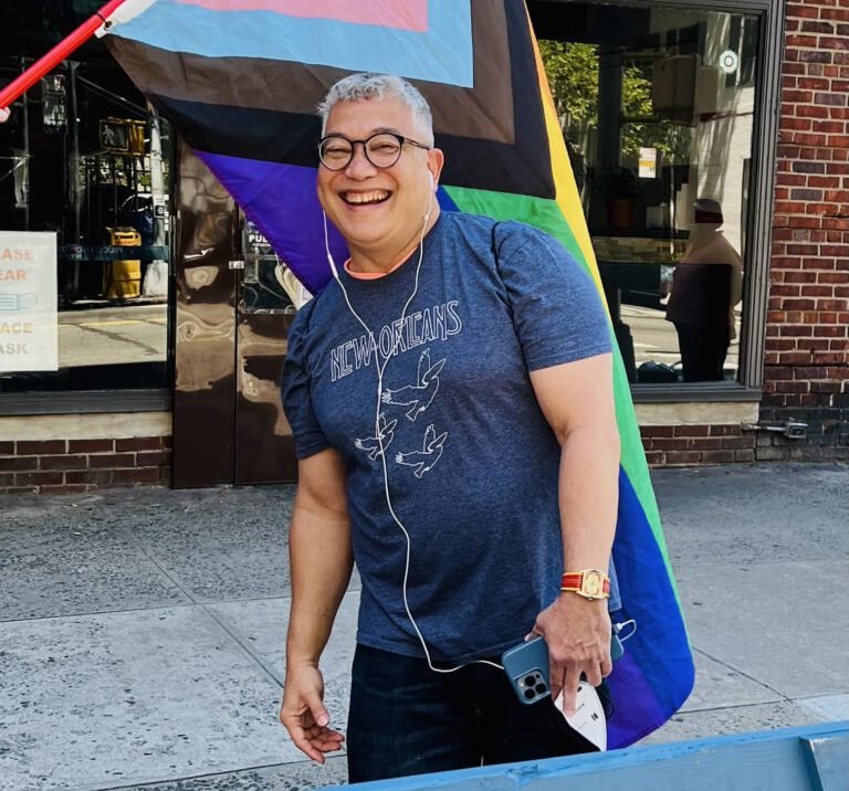 A person with glasses smiling in front of an LGBTQ+ pride flag.