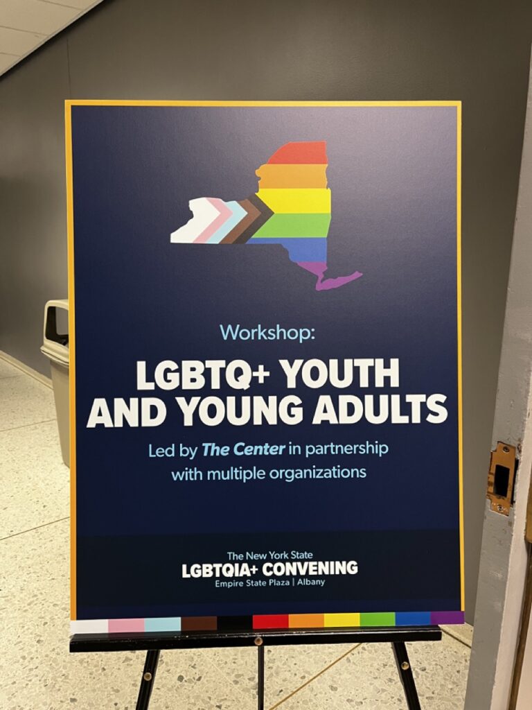 a dark blue sign that reads Workshop LGBTQ+ Youth And Young Adults with an outline of New York State filled in with progress flag colors