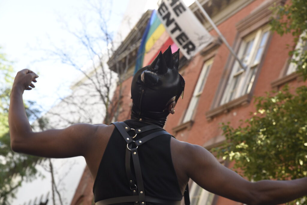 a Black person wearing black leather tank top and headpiece stands with arms open an their back to you as the pride flag flies over them. The Center is on the flag in black letters