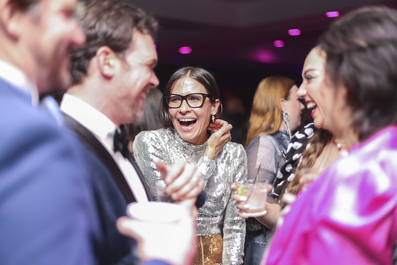 a dark haired femme presenting person wearing dark rimmed glasses smiles at as others look on in revelry. All are dressed in festive evening wear.