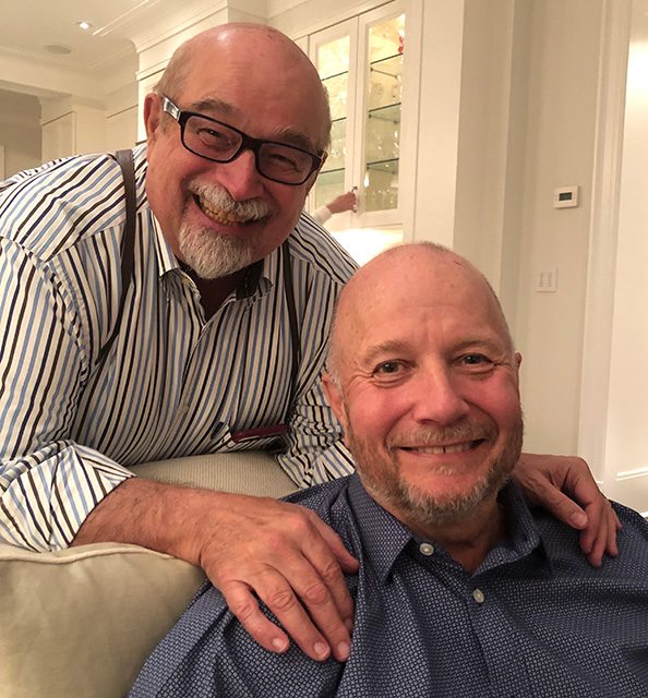 two white men smiling at you. one is sitting on the couch wearing a blue shirt, the over is behind the couch with his hands on the other man's shoulders. He's wearing a striped shirt and glasses. Both have short beards and are bald