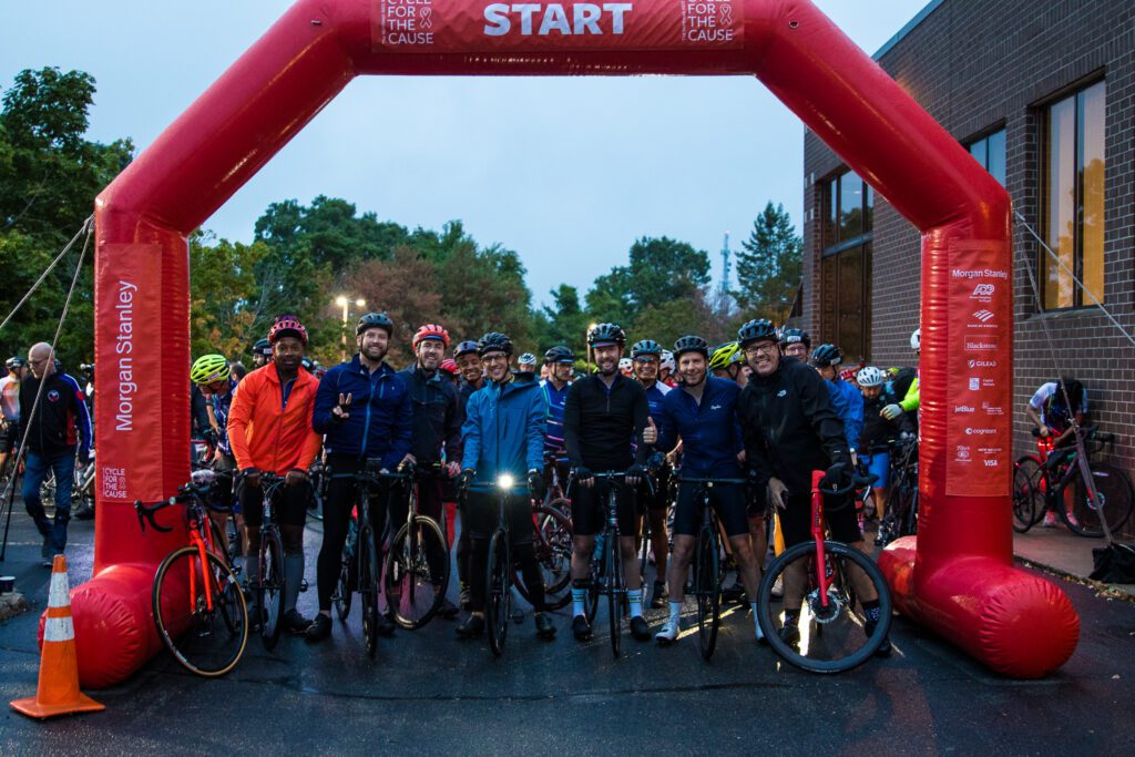 A group of cyclists stand with bikes under red ballon arch with Start written on it