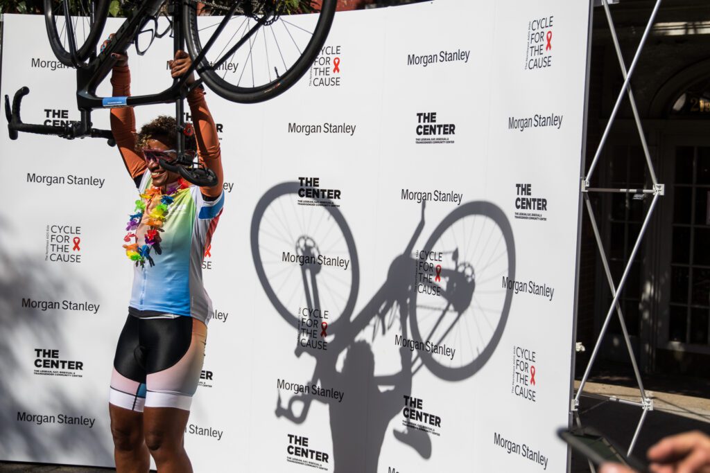 a women lifts her bike over her head celebrating.she is wearing a cyclist uniform and standing in front of a backdrop with The Center, Cycle for the Cause, and Morgan Stanley logos