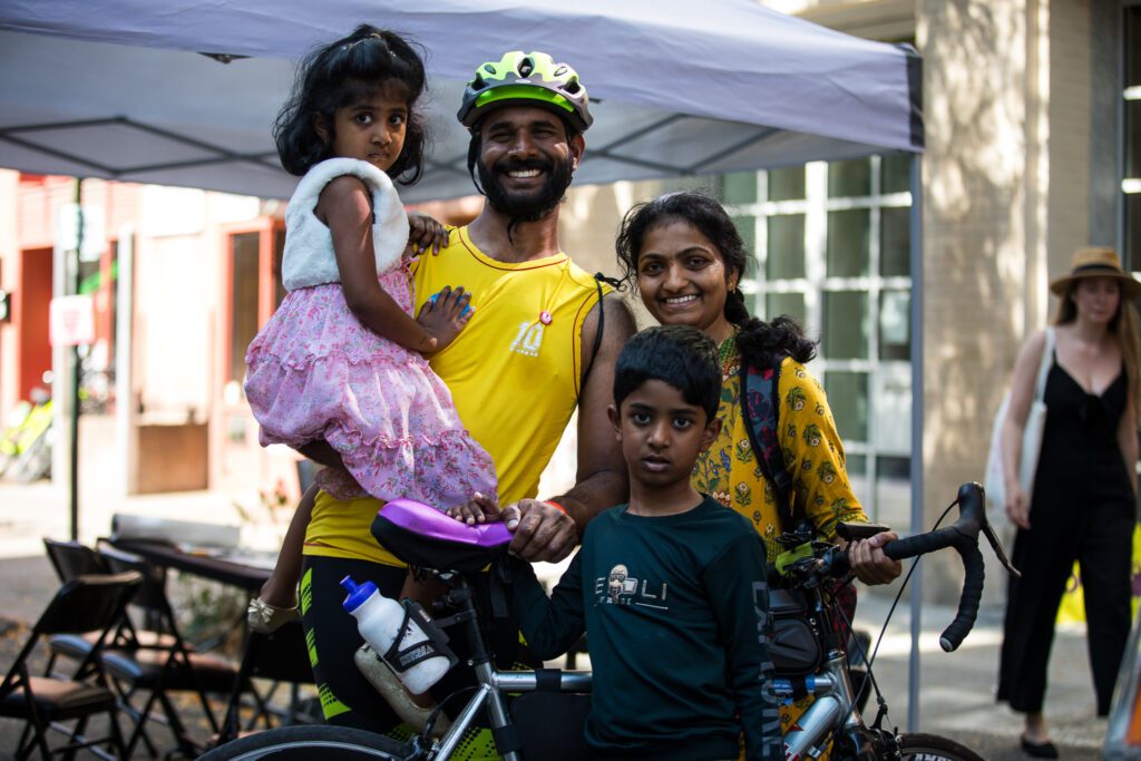 an family poses with a father figure who is a cyclist, a mother, and two kids. The father is holding the smaller child, who is wearing a pink and white dress. They have brown skin.