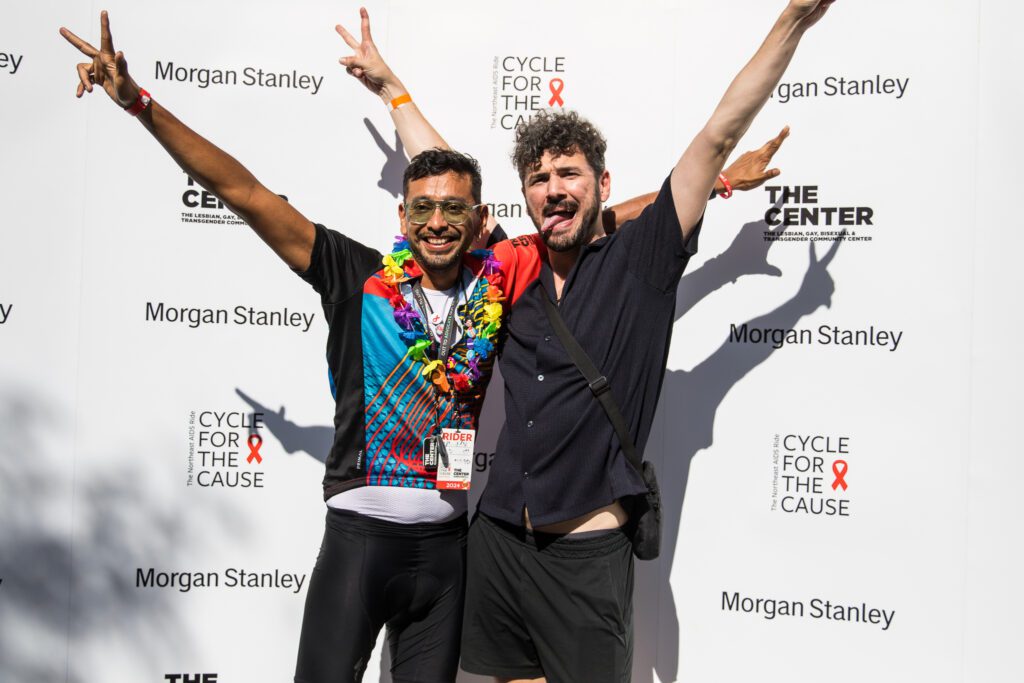 two masc presenting people celebrate with arms in the air as they stand in front of a backdrop with the Cycle for the Cause and Morgan Stanely logos on it