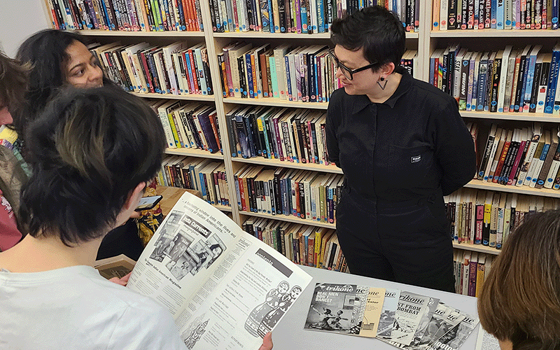 person standing in front of a bookshelf smiling with a group of youth who are looking at an old magazine