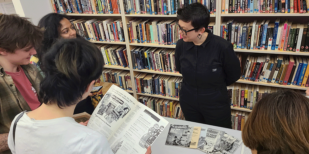 person standing in front of a bookshelf smiling with a group of youth who are looking at an old magazine