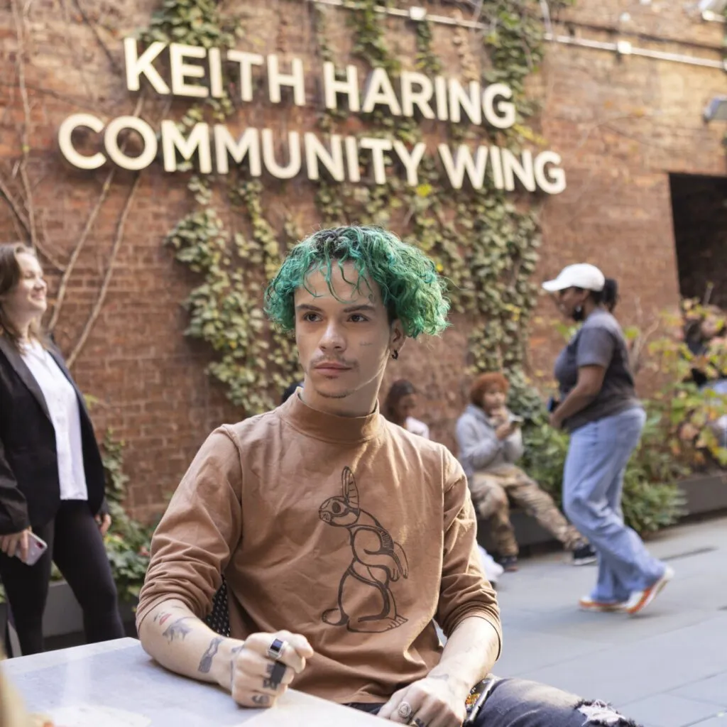 A young person with vibrant green hair sits outside under the Keith Haring Community Wing at The Center