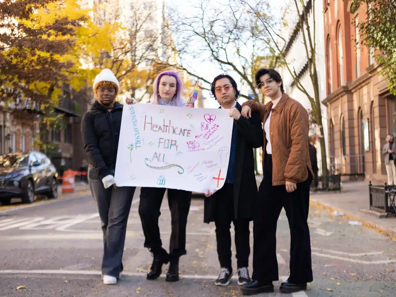 Four people of multiple gender identities, standing on a New York City street holding a sign advocating for better health care & HIV and AIDS