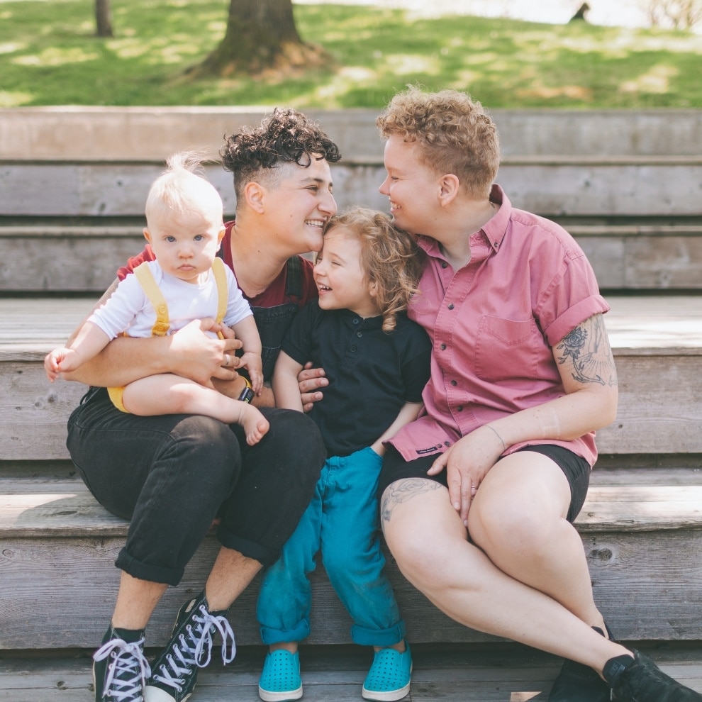 Two women sitting closely with two children. They are smiling and are outdoors seated on stone steps like one would see at a park