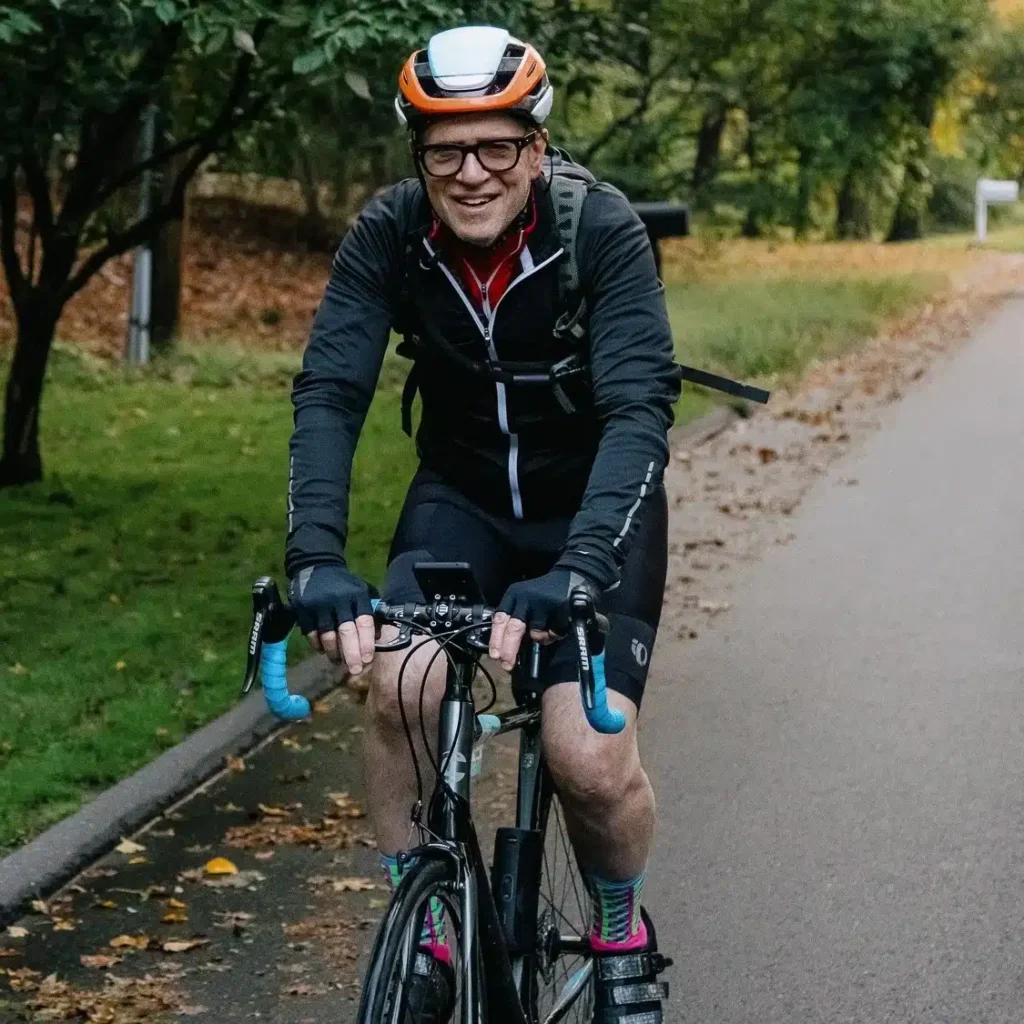 A cyclist wearing an orange and white helmet, glasses, and a black cycling jacket rides a road bike along a tree-lined path. The rider is smiling, wearing gloves and colorful socks, with autumn leaves scattered on the ground.